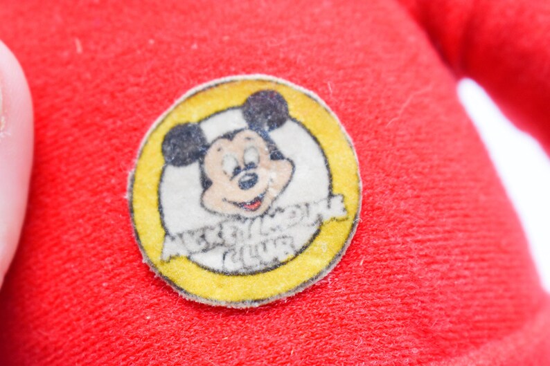 May include: Close-up of a red fabric with a circular Mickey Mouse Club patch. The patch features a smiling Mickey Mouse in a yellow circle with the words "Mickey Mouse Club".