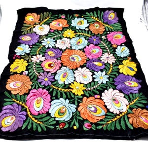 May include: Black textile with vibrant floral embroidery. The design features a central arrangement of colorful flowers in shades of orange, pink, purple, and blue, surrounded by green leaves and a yellow border.