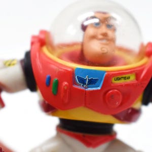 May include: A close-up of a Buzz Lightyear action figure. The figure has a red and white spacesuit with a clear helmet. The chest features a blue emblem with a flying bird design and the word "LIGHTYEAR".