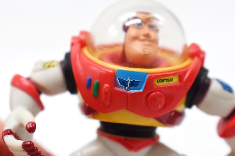 May include: A close-up of a Buzz Lightyear action figure. The figure has a red and white spacesuit with a clear helmet. The chest features a blue emblem with a flying bird design and the word "LIGHTYEAR".