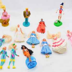 Op de afbeelding: Een verzameling vintage miniatuurpoppen, waaronder figuren in jurken, sportkleding en formele kleding. De poppen zijn in verschillende kleuren, met blond en donker haar. Sommige poppen staan op voetstukken.