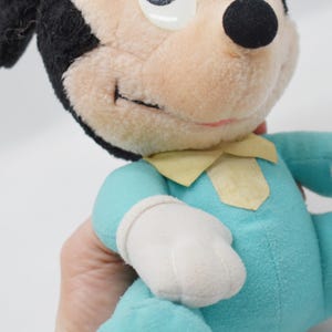 Op de afbeelding: Een vintage Mickey Mouse pluche speelgoed. Het pluche heeft een zwarte kop en oren, een lichtbruin gezicht en een lichtblauwe body met witte handschoenen. Het heeft een gele kraag en een gestikte glimlach.