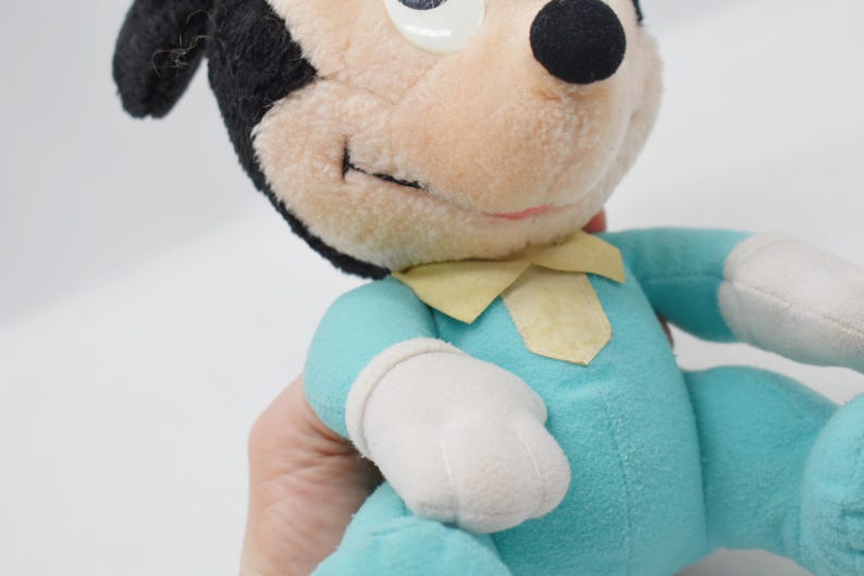 Op de afbeelding: Een vintage Mickey Mouse pluche speelgoed. Het pluche heeft een zwarte kop en oren, een lichtbruin gezicht en een lichtblauwe body met witte handschoenen. Het heeft een gele kraag en een gestikte glimlach.