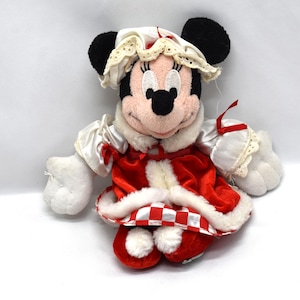May include: A plush Minnie Mouse doll wearing a red and white outfit with a matching bonnet. The doll has black ears, a pink face, and white gloves. The outfit includes a red and white checkered trim.