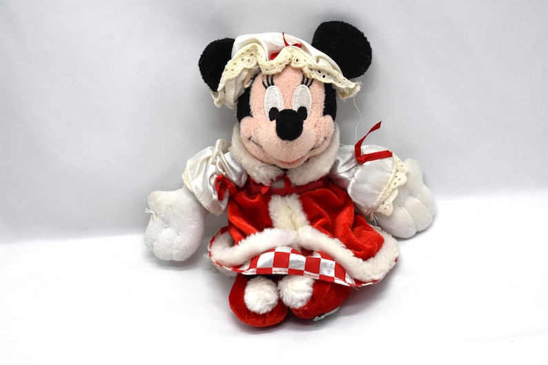 May include: A plush Minnie Mouse doll wearing a red and white outfit with a matching bonnet. The doll has black ears, a pink face, and white gloves. The outfit includes a red and white checkered trim.