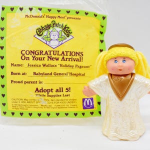 May include: A Cabbage Patch Kids doll from a McDonald's Happy Meal. The doll has blonde hair, blue eyes, and is wearing a white and gold outfit. The packaging includes text that reads "CONGRATULATIONS On Your New Arrival!" and "Adopt all 5!"