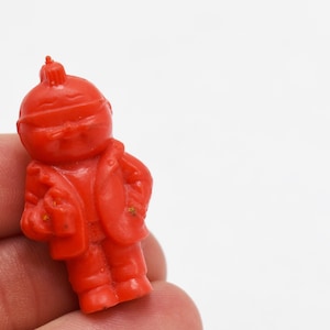 May include: A small, vintage, red plastic figurine of a person wearing a hat and jacket. The figurine has a detailed design and is held in a hand against a white background. The figurine is a collectible toy.