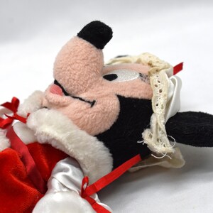 May include: A plush toy featuring a character with a pink face, black ears, and a black nose. The toy is dressed in a red velvet outfit with white trim and red ribbons. A white lace-trimmed bonnet is on the character's head.