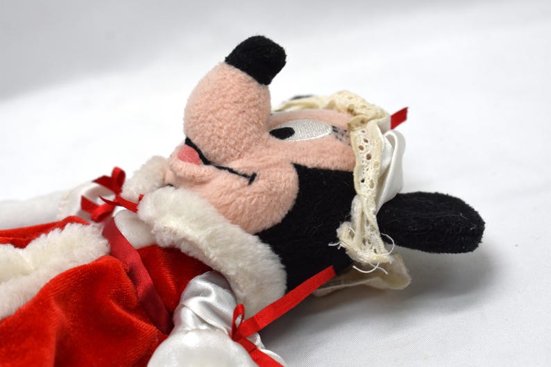 May include: A plush toy featuring a character with a pink face, black ears, and a black nose. The toy is dressed in a red velvet outfit with white trim and red ribbons. A white lace-trimmed bonnet is on the character's head.