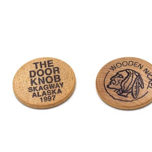 Puede incluir: Dos fichas redondas de madera con impresión en tinta negra. Una ficha muestra el texto THE DOOR KNOB SKAGWAY ALASKA 1997. La otra ficha presenta una ilustración de perfil de una persona con un tocado y el texto WOODEN NICKEL impreso alrededor.