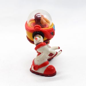 May include: A vintage toy robot with a clear dome head, red and yellow body, and white and red legs. The robot has a retro design, with a small figure visible inside the dome. The toy is likely from the 1980s or 1990s.