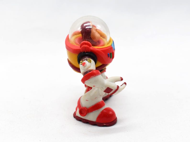 May include: A vintage toy robot with a clear dome head, red and yellow body, and white and red legs. The robot has a retro design, with a small figure visible inside the dome. The toy is likely from the 1980s or 1990s.