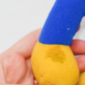 May include: A plush toy's blue arm and yellow foot are visible. The toy is held in a person's hand. The foot has a yellow stain. The background is white.