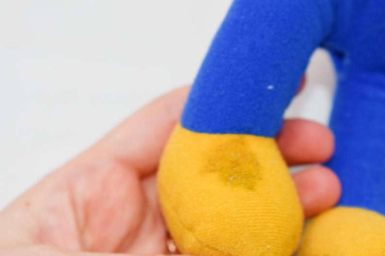 May include: A plush toy's blue arm and yellow foot are visible. The toy is held in a person's hand. The foot has a yellow stain. The background is white.