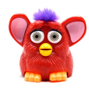 May include: A red electronic toy creature with a purple tuft of hair on its head, large white eyes with black radial iris details, prominent red ears with pink inner sections, a yellow beak, and orange feet, standing against a white background.