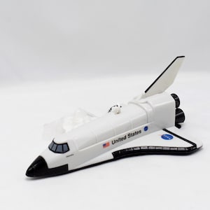 May include: A white toy space shuttle with black accents, including the nose cone, engine nozzles, and wing edges. The words "United States" and a US flag are printed on the side. The NASA logo is also visible.