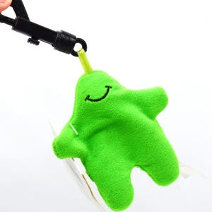 May include: A bright green plush keychain in the shape of a smiling blob. The keychain has a black clip and a small yellow loop connecting it to the plush toy. The toy has a stitched black smile.