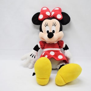 K&ouml;nnte beinhalten: Ein Pl&uuml;schtier von Minnie Mouse, die ein rotes Kleid mit wei&szlig;en Punkten und eine rote Schleife mit wei&szlig;en Punkten tr&auml;gt. Sie hat schwarze Augen, eine schwarze Nase und gelbe Schuhe.