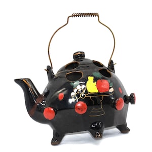 Puede incluir: Un objeto decorativo de cerámica negra con forma de tetera con un asa de alambre. La tetera presenta lunares rojos y un diseño de frutas pintadas. El pico y el asa son marrones. Estética vintage única.