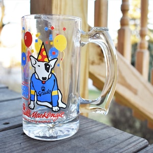 May include: Clear glass beer mug featuring a cartoon Bull Terrier wearing a blue sweater and party hat. The mug has a handle and is decorated with balloons and the text "Spuds Mackenzie".