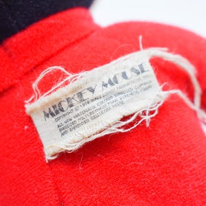 May include: Close-up of a vintage Mickey Mouse plush toy. The toy is primarily red with black accents. A rectangular tag with the words "Mickey Mouse" is sewn onto the plush, with additional text below.