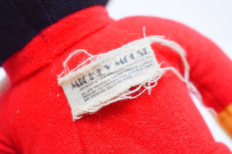 May include: Close-up of a vintage Mickey Mouse plush toy. The toy is primarily red with black accents. A rectangular tag with the words "Mickey Mouse" is sewn onto the plush, with additional text below.