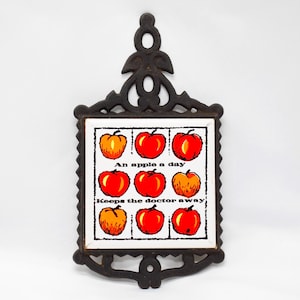 May include: A cast iron trivet with a decorative frame and a ceramic tile insert. The tile features nine illustrated apples in red and orange, with the text "An apple a day keeps the doctor away."