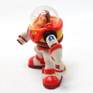 May include: A vintage astronaut action figure in a red, yellow, and white spacesuit with a clear helmet. The figure has a detailed face and is posed as if walking. The spacesuit has a blue star emblem and the word "OPEN" on the chest.