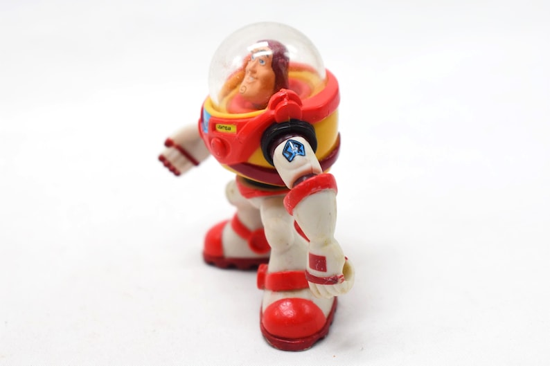 May include: A vintage astronaut action figure in a red, yellow, and white spacesuit with a clear helmet. The figure has a detailed face and is posed as if walking. The spacesuit has a blue star emblem and the word "OPEN" on the chest.