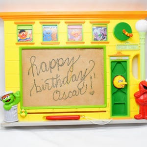 May include: A yellow Sesame Street-themed drawing board with a green screen and the words "happy birthday Oscar!" written on it. The board features images of Sesame Street characters, including Elmo, Big Bird, and Oscar the Grouch.