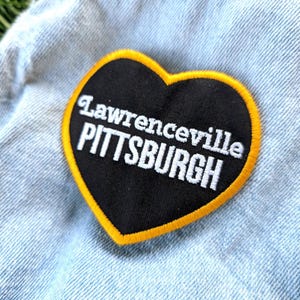 May include: A heart-shaped patch with the text "Lawrenceville PITTSBURGH" in white on a black background, outlined in yellow. The patch is sewn onto a light blue denim fabric.