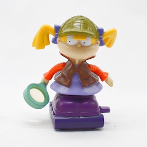 Puede incluir: Figura de juguete colorida de un personaje de dibujos animados con coletas amarillas, que lleva un sombrero verde, un chaleco marrón, mangas naranjas y un vestido morado. La figura sostiene una lupa y está montada sobre una base morada.