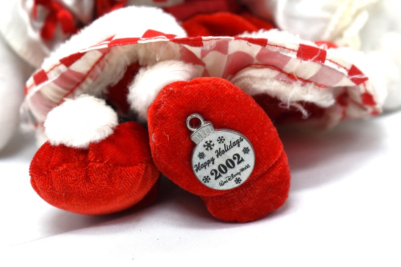 May include: Close-up of a plush toy's red foot with a white pom-pom and a silver ornament. The ornament reads "Happy Holidays 2002" and features snowflakes. The toy has a red and white checkered pattern.