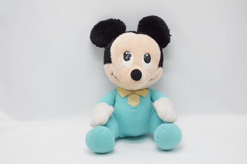 Op de afbeelding: Een pluche Mickey Mouse pop, met zwarte oren, een bruin gezicht en een turquoise outfit met een gele vlinderdas. De pop heeft witte handschoenen en turquoise voeten. Het speelgoed zit op een witte achtergrond.