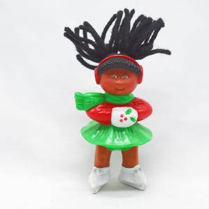Op de afbeelding: Vintage poppenbeeldje met zwart garen haar, draagt een rode hoofdband, sjaal en top. De pop heeft een groene rok, witte wanten met hulstbessen en witte schaatsen. Kerstversiering.