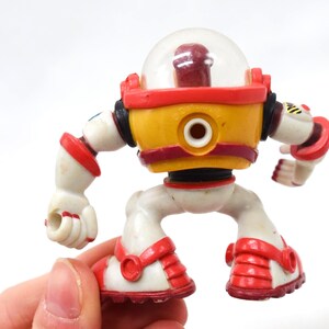 May include: A vintage toy robot with a clear dome head, yellow torso, and red accents. The robot has white limbs and red details on its feet and arms. The toy is held in a hand against a white background.