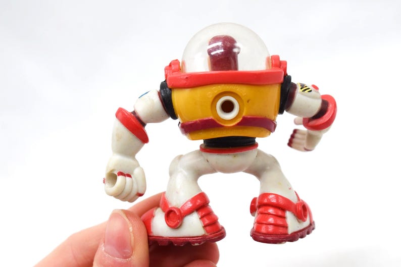 May include: A vintage toy robot with a clear dome head, yellow torso, and red accents. The robot has white limbs and red details on its feet and arms. The toy is held in a hand against a white background.