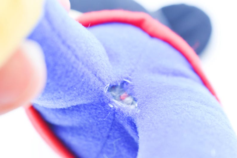 May include: Close-up of a torn blue plush toy with red and black accents. The tear reveals the inner stuffing of the toy. A hand is partially visible in the image.