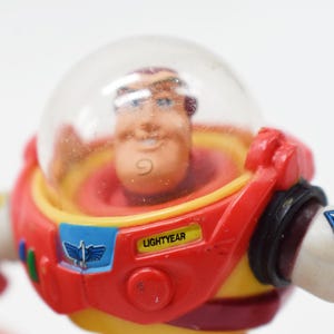 May include: A close-up of a Buzz Lightyear action figure. The figure has a clear plastic helmet, a red and yellow spacesuit, and the name "LIGHTYEAR" printed on the chest. The figure is a collectible toy.
