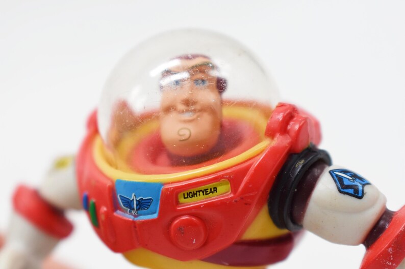 May include: A close-up of a Buzz Lightyear action figure. The figure has a clear plastic helmet, a red and yellow spacesuit, and the name "LIGHTYEAR" printed on the chest. The figure is a collectible toy.