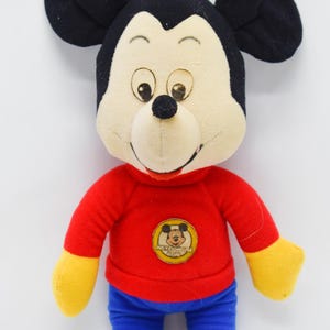 May include: A plush Mickey Mouse doll with a red shirt, blue pants, and yellow gloves. The doll has black ears, a cream-colored face, and a small black nose. A Mickey Mouse emblem is on the shirt.