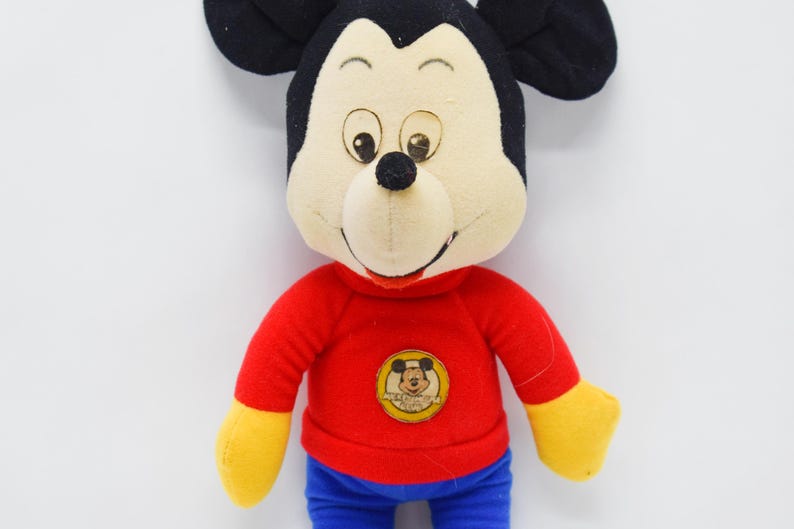May include: A plush Mickey Mouse doll with a red shirt, blue pants, and yellow gloves. The doll has black ears, a cream-colored face, and a small black nose. A Mickey Mouse emblem is on the shirt.