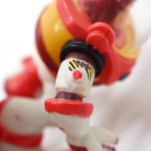 May include: A vintage astronaut toy in red, yellow, and white. The toy has a clear helmet, black accents, and a red nose. The astronaut's arm is bent, and the toy is set against a white background.