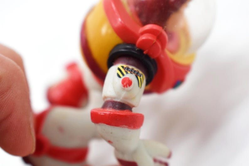 May include: A vintage astronaut toy in red, yellow, and white. The toy has a clear helmet, black accents, and a red nose. The astronaut's arm is bent, and the toy is set against a white background.