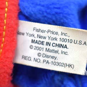 K&ouml;nnte beinhalten: Nahaufnahme eines Etiketts von einem Pl&uuml;schtier, mit rotem und blauem Stoff. Auf dem Etikett steht "Fisher-Price, Inc., New York, New York 10010 U.S.A. MADE IN CHINA. &copy;2001 Mattel, Inc. &copy;Disney. REG. NO. PA-10302(HK)".