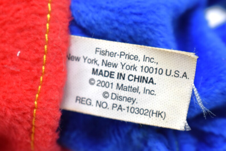 K&ouml;nnte beinhalten: Nahaufnahme eines Etiketts von einem Pl&uuml;schtier, mit rotem und blauem Stoff. Auf dem Etikett steht "Fisher-Price, Inc., New York, New York 10010 U.S.A. MADE IN CHINA. &copy;2001 Mattel, Inc. &copy;Disney. REG. NO. PA-10302(HK)".