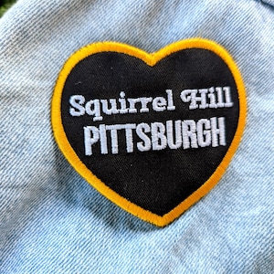 May include: A heart-shaped patch with the words "Squirrel Hill PITTSBURGH" in white lettering on a black background. The patch has a yellow border and is set against a light blue denim fabric.