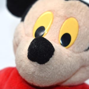 K&ouml;nnte beinhalten: Nahaufnahme eines Mickey Mouse-Pl&uuml;schtiers. Das Spielzeug hat eine schwarze Nase, schwarze Ohren und gelbe Augen mit schwarzen Pupillen. Das Pl&uuml;schtier tr&auml;gt ein rotes Hemd. Der Hintergrund ist wei&szlig;.