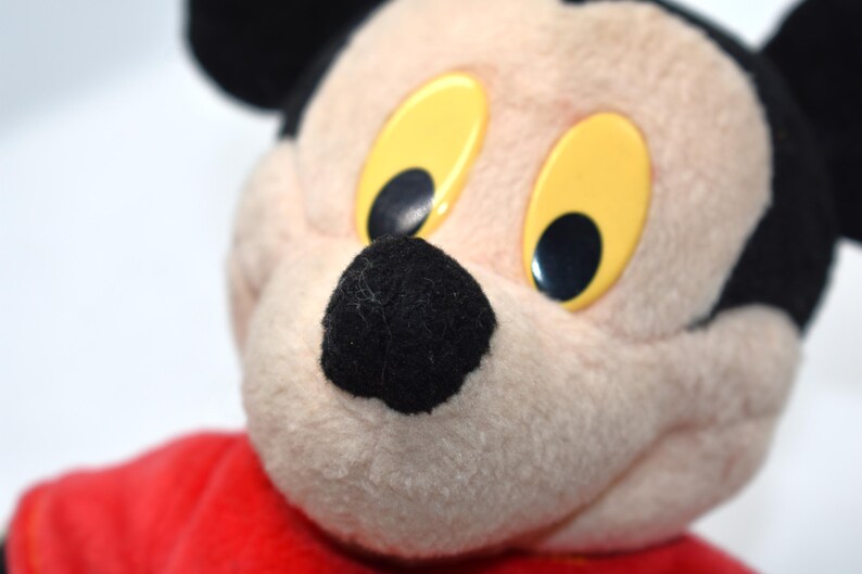 K&ouml;nnte beinhalten: Nahaufnahme eines Mickey Mouse-Pl&uuml;schtiers. Das Spielzeug hat eine schwarze Nase, schwarze Ohren und gelbe Augen mit schwarzen Pupillen. Das Pl&uuml;schtier tr&auml;gt ein rotes Hemd. Der Hintergrund ist wei&szlig;.