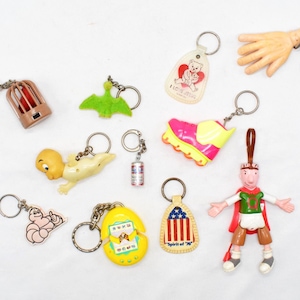 May include: A collection of vintage keychains with various designs, including a bird in a cage, a green pterodactyl, a baby, a miniature can, a running shoe, a hand, a bear with a heart, a worm, a cracked egg, and a patriotic design.